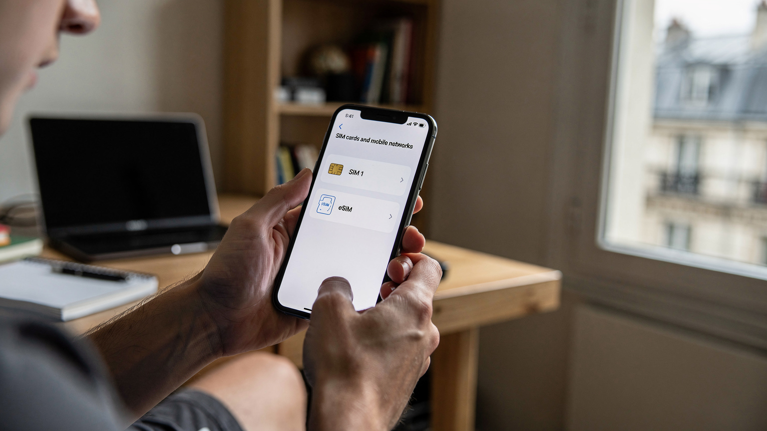 En ung student holder en smarttelefon og viser innstillingsmenyen der to SIM-kort, ett fysisk og ett eSIM, er aktive, i en studentbolig i Paris.