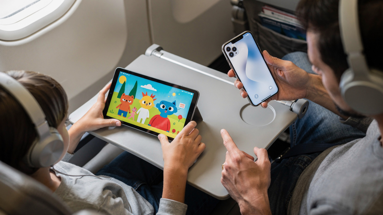 Mãos de um adulto e uma criança sentadas em um avião, com a criança assistindo a desenhos animados em um tablet e o adulto verificando a conexão em um iPhone 17, ambos com fones de ouvido.