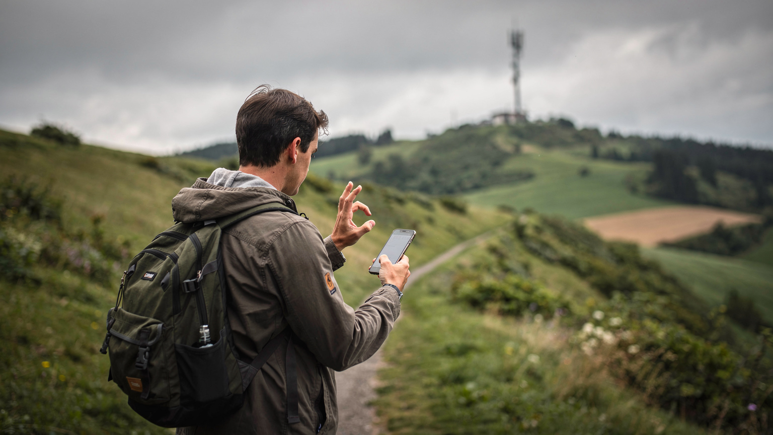 Eine Person versucht, in einem ländlichen Gebiet auf einer Karte auf ihrem Smartphone nach Empfang zu suchen, während der Hintergrund unscharfe grüne Hügel und einen Sendemast zeigt.