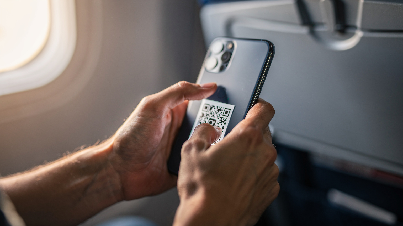 Close-up of a traveler's hands holding a smartphone, scanning a QR code for eSIM activation, with a blurred airplane window in the background.