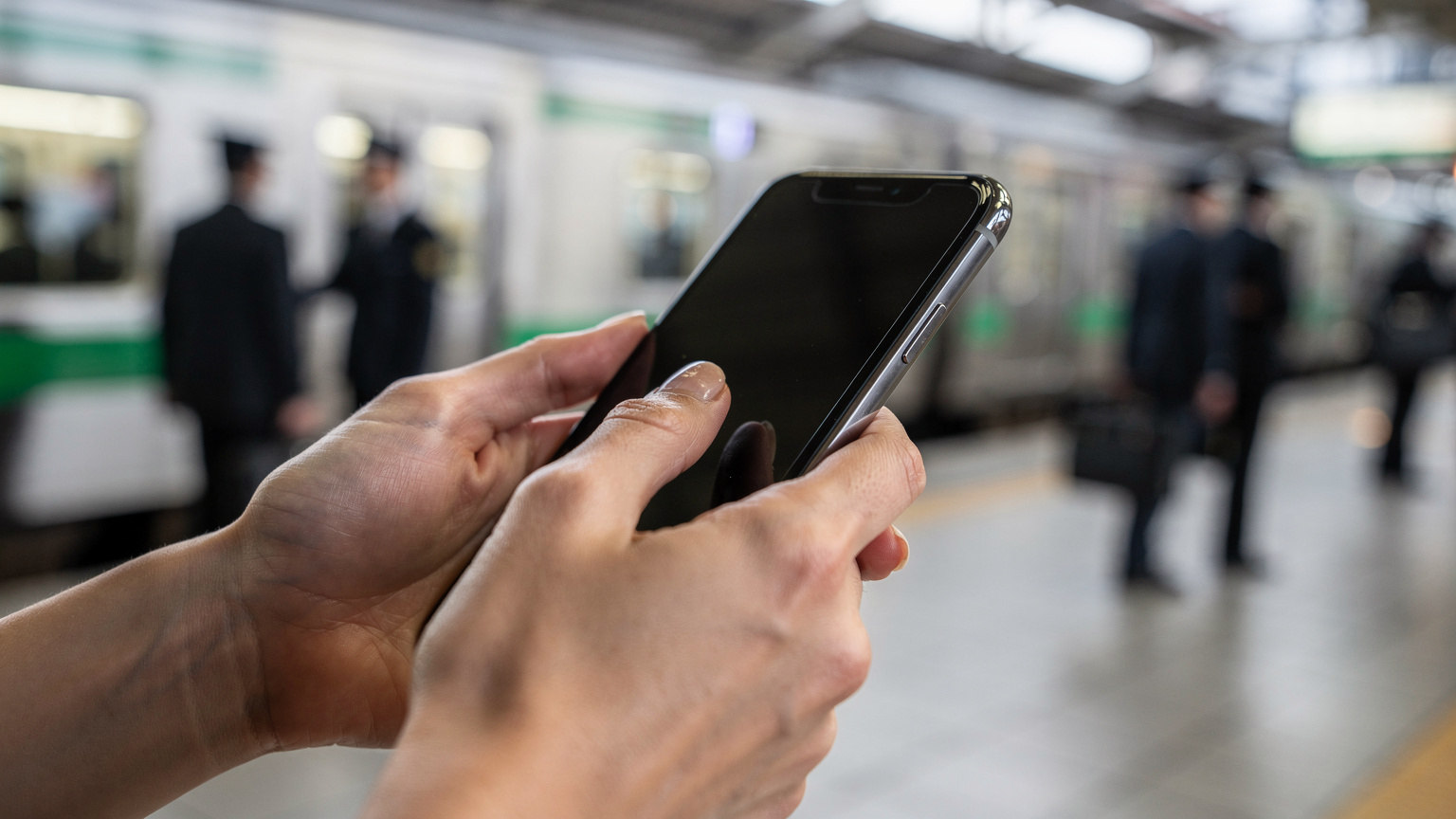 Zblízka detail ženských rúk, ktoré držia smartfón a prechádzajú prstom po obrazovke, s rozostreným pozadím rušného vlakového nástupišťa v Tokiu (napríklad stanica Yamanote).