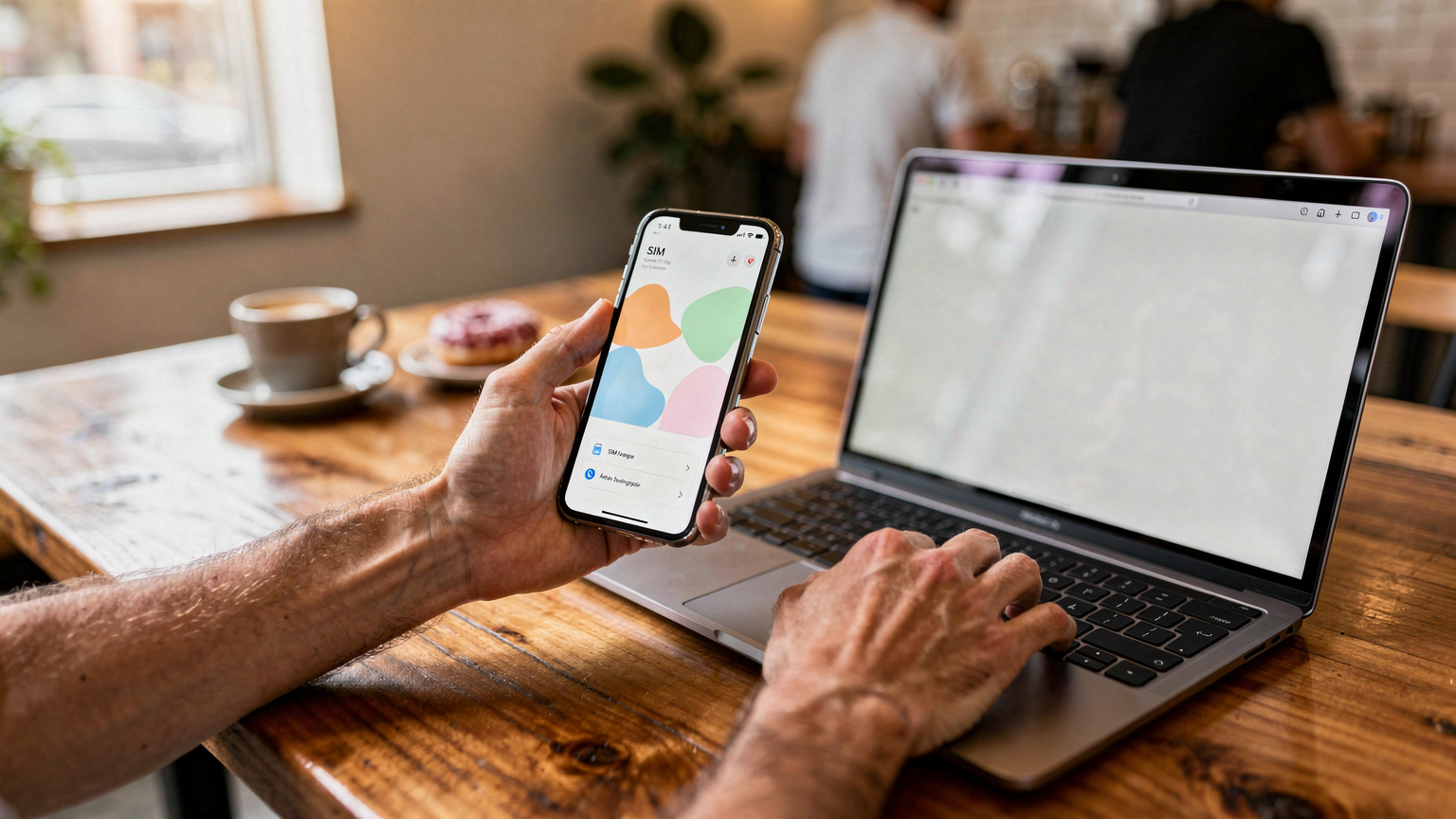 Ruce digitálního nomáda v kavárně v Seattlu, jak drží svůj smartphone s otevřenou aplikací pro správu eSIM a zároveň píše na notebooku. V pozadí je rozmazaný interiér kavárny s dřevěnými stoly a dalšími lidmi.