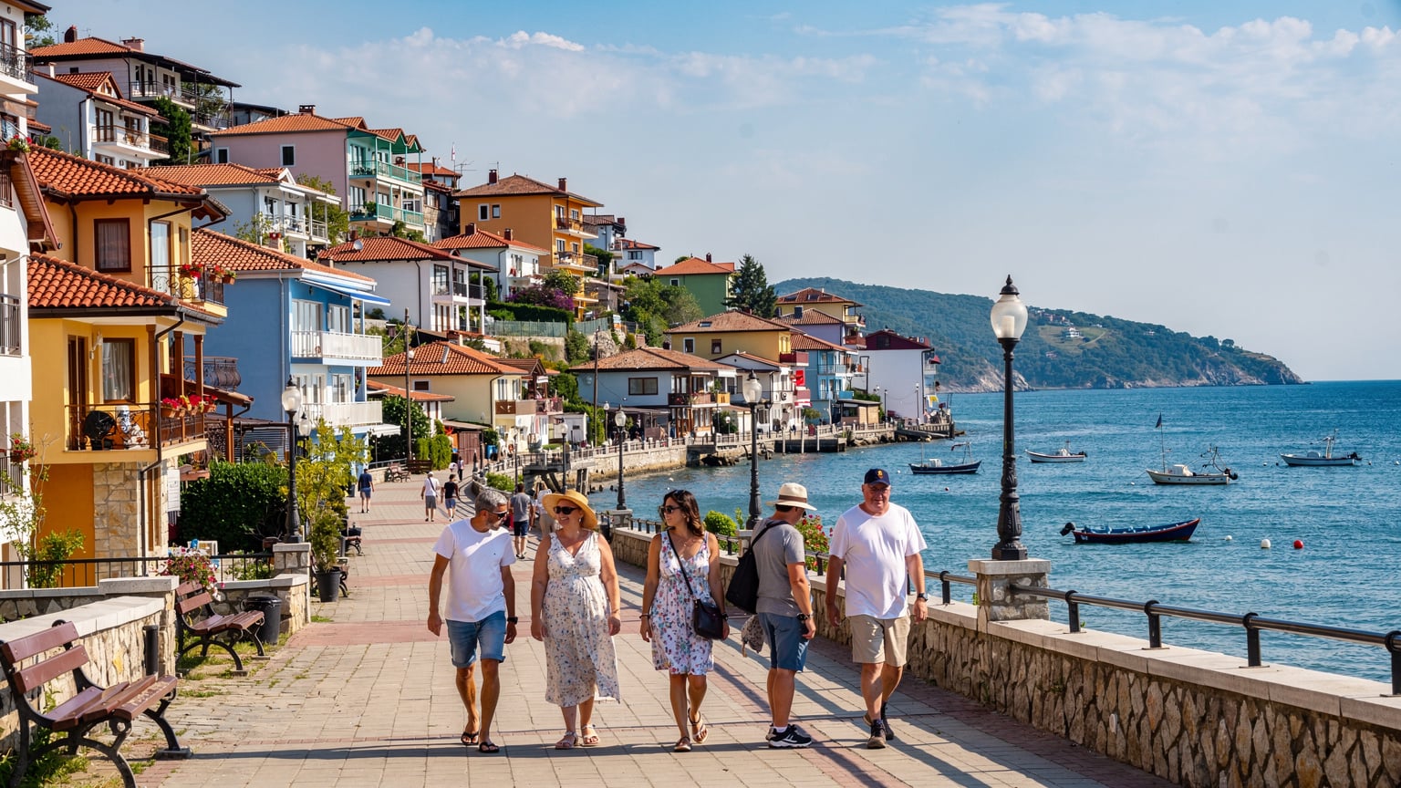 Un grup de turiști se plimbă pe o promenadă pietruită de-a lungul coastei, în fața unor case colorate și pitorești din Balchik, Bulgaria, cu Marea Neagră calmă în fundal și bărci de pescuit la orizont.