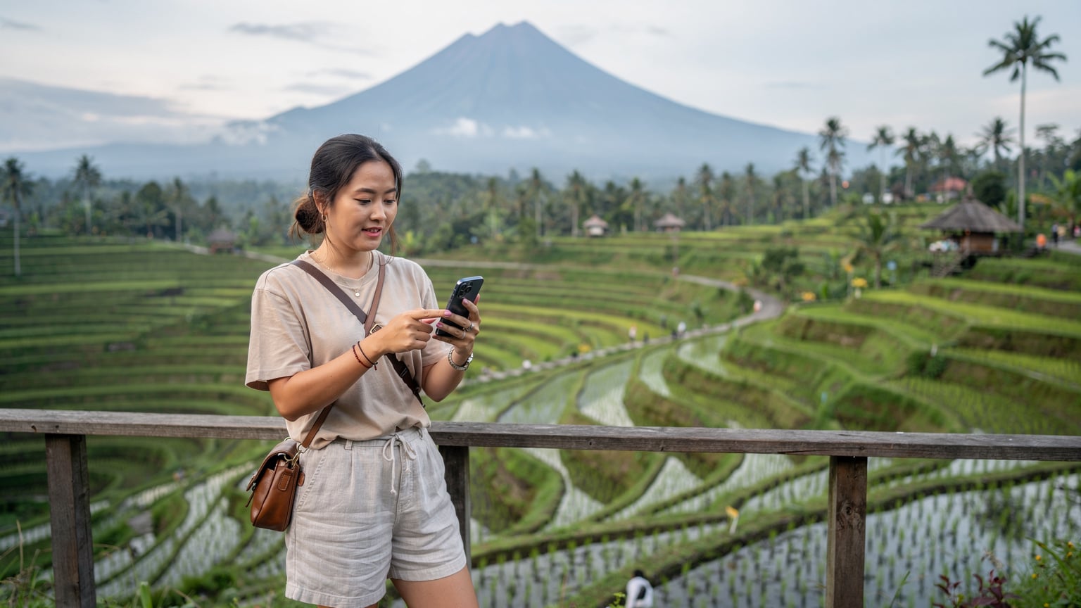 Seorang wanita Asia yang sedang melakukan perjalanan memegang ponselnya dan memindai kode QR eSIM dengan latar belakang sawah hijau terasering dan gunung berapi di kejauhan di Bali.
