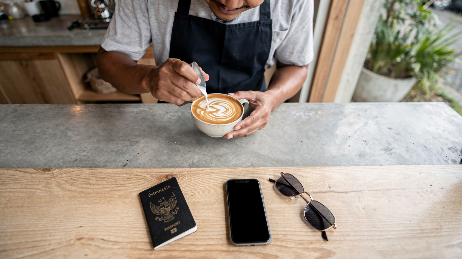 Seorang barista di kafe trendi di Canggu sedang menyajikan kopi, sementara di latar depan ada ponsel, paspor Indonesia, dan kacamata hitam di meja.