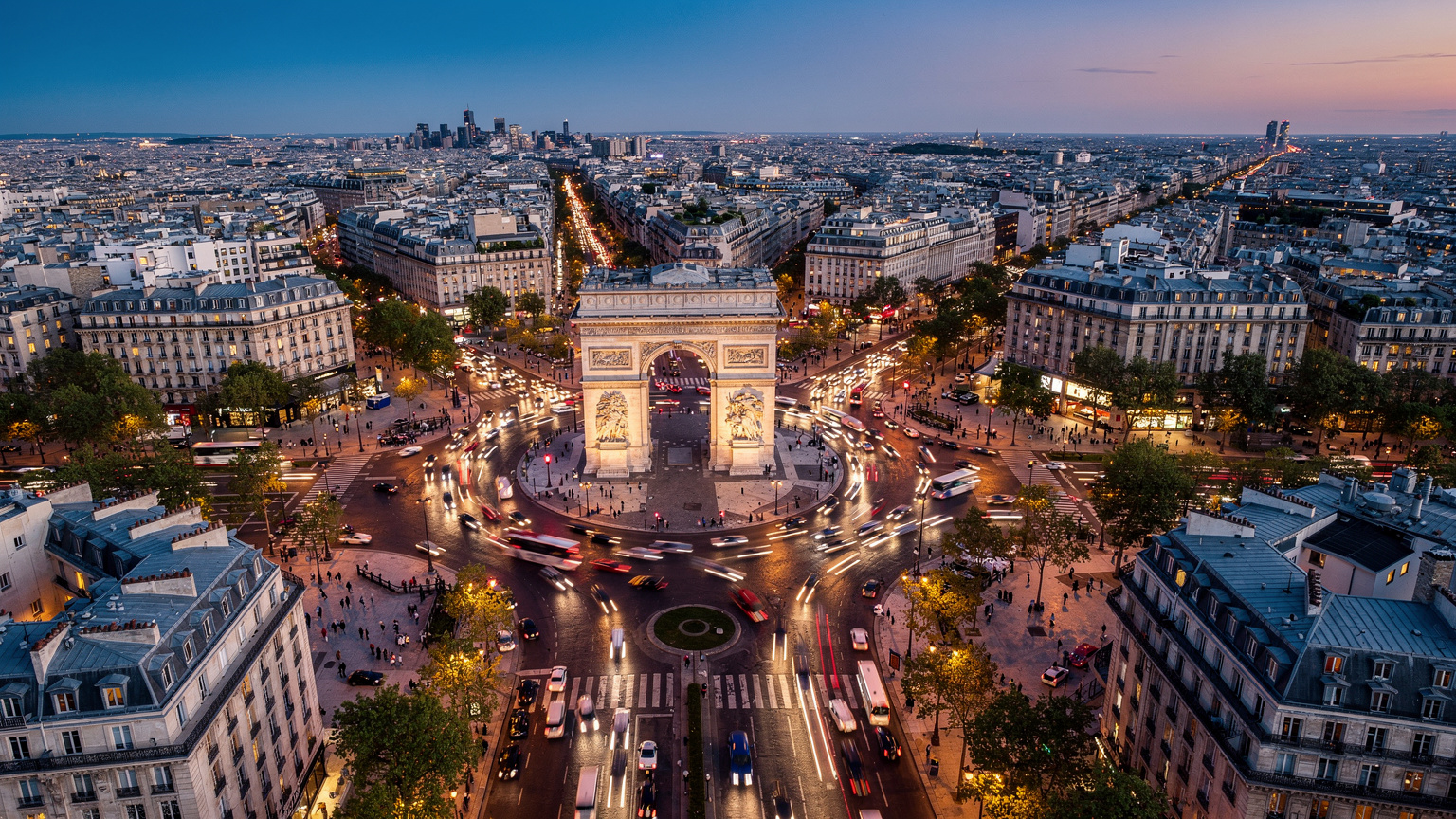 Vue aérienne de l'Arc de Triomphe et de la Place Charles de Gaulle, avec des avenues rayonnantes remplies de voitures et de piétons, au crépuscule, sous des lumières de ville scintillantes.