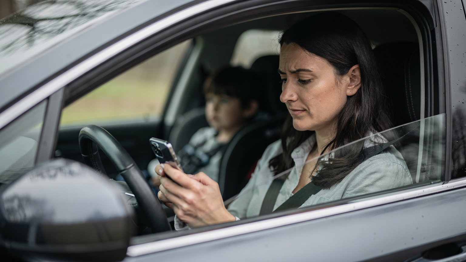 A worried parent in a car, holding their smartphone and looking at the screen with a slight frown, indicating a connectivity issue, while a child watches from the back seat.