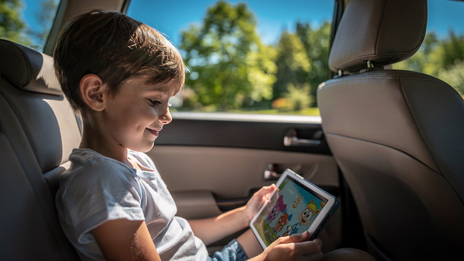 A child in the back seat of a car happily watching a show on a tablet, with a bright summer landscape visible through the window.