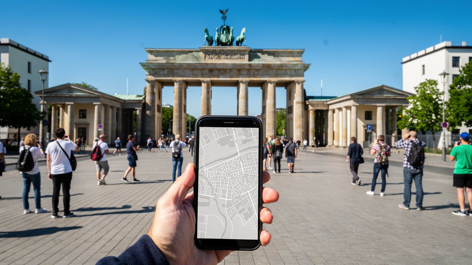 Ein Reisender hält sein Smartphone mit einer geöffneten Karten-App in der Hand, während er vor dem Brandenburger Tor steht. Im Hintergrund sind andere Touristen und das historische Bauwerk zu sehen.