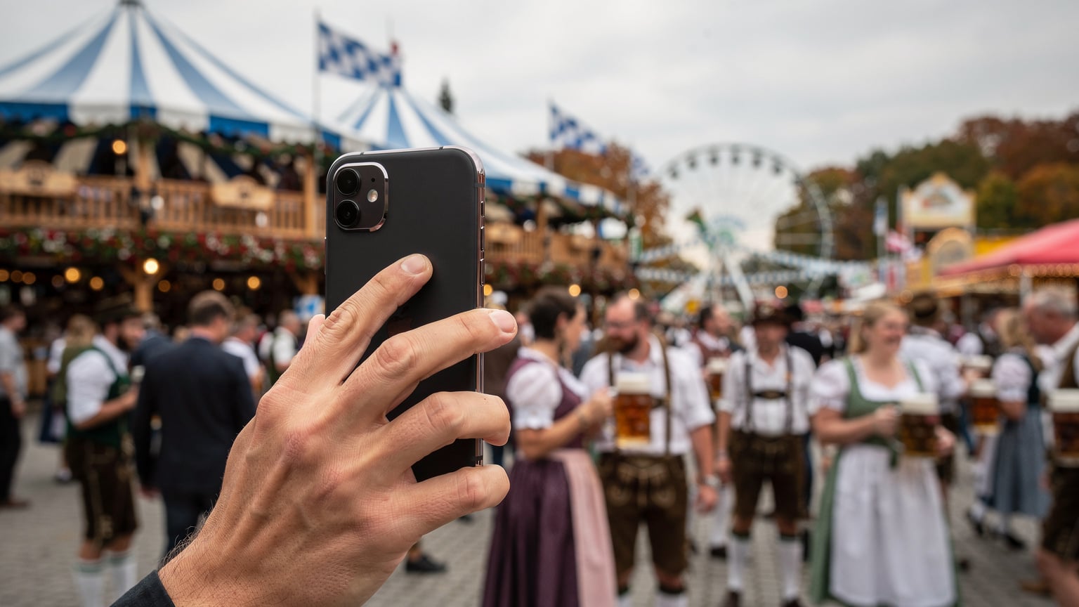 A person's hand holding a smartphone, capturing the lively atmosphere of Oktoberfest with beer tents and festive crowds in the background.