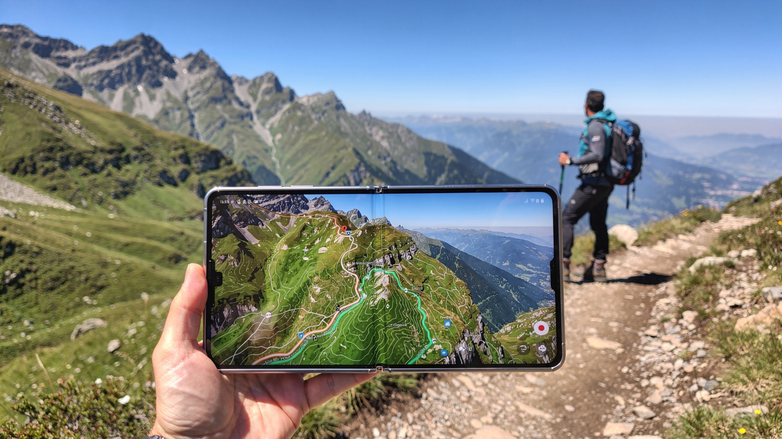 Un Samsung Galaxy Z Fold 8 desplegado mostrando una aplicación de mapas con la ruta de un sendero de montaña en los Pirineos, con una persona con equipo de senderismo en segundo plano, disfrutando de la conexión en la naturaleza.