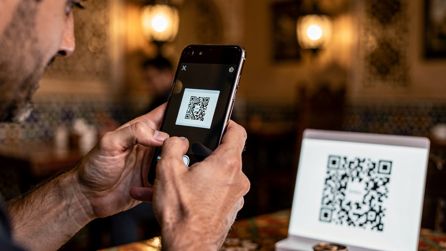 Close-up of human hands holding a smartphone, showing the action of scanning a QR code for eSIM activation, with a blurred Moroccan cafe background.