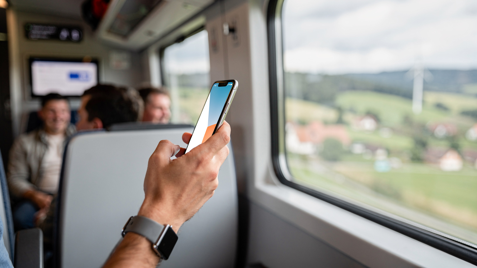 Рука молодого мандрівника в поїзді Німецьких залізниць (Deutsche Bahn), обирає план eSIM на екрані телефону, з видом на сільські краєвиди за вікном.