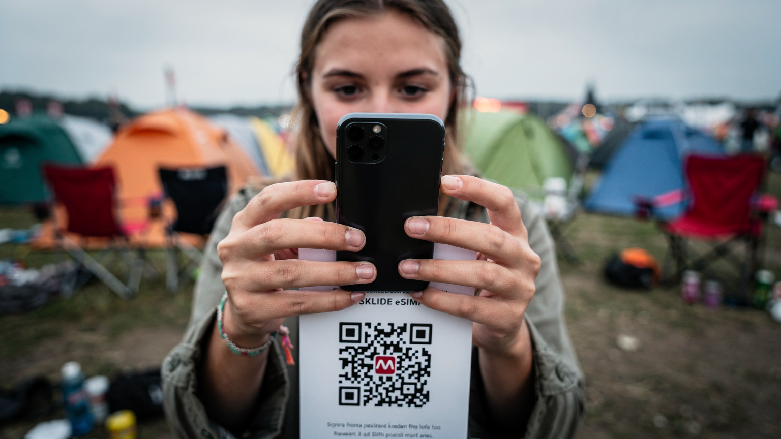 En ung kvinde scanner en QR-kode med sin smartphone for at aktivere et eSIM, hendes campingudstyr er synligt i baggrunden af festivalområdet.