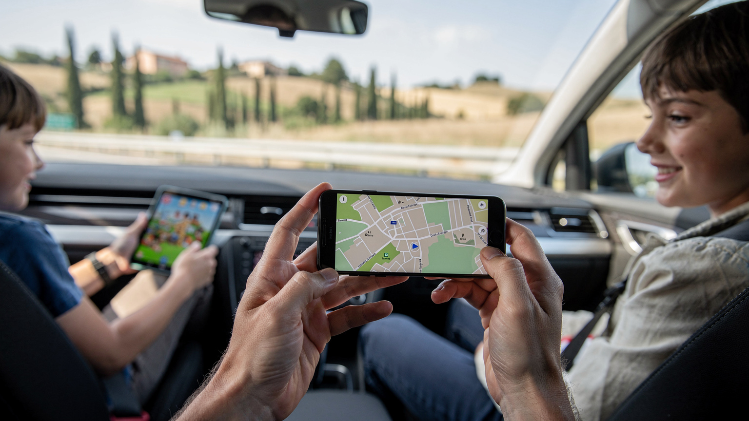 Mani di un adulto con smartphone che mostra Google Maps, mentre i bambini guardano un tablet nei sedili posteriori di un'auto in autostrada in Italia.