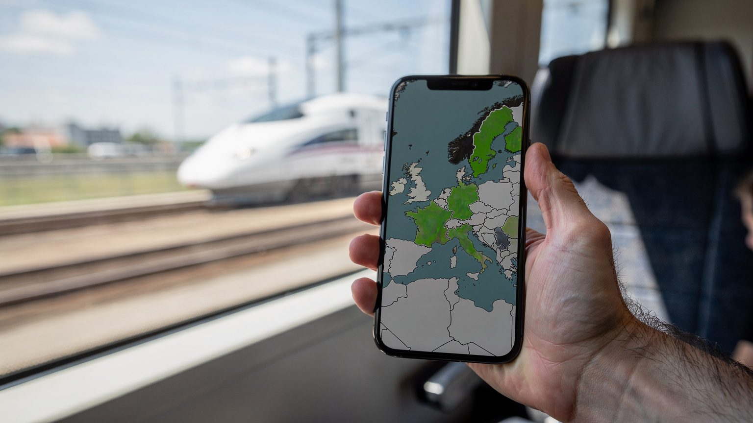 Un smartphone affichant une carte de couverture réseau avec des zones vertes (bonne couverture) et des zones grises (faible couverture) est tenu par une main masculine, avec un train à grande vitesse flou en arrière-plan, évoquant le voyage rapide en Europe.