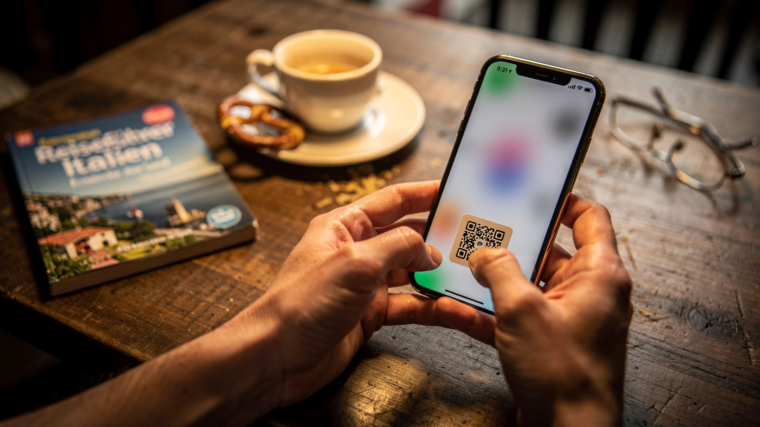 Nahaufnahme von Händen, die ein Smartphone halten und den QR-Code einer eSIM scannen, während im Hintergrund ein Reiseführer und eine Tasse Kaffee auf einem Tisch liegen.