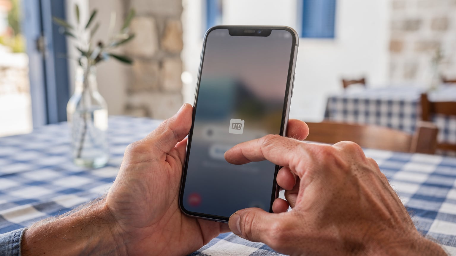 A close-up shot of a traveler's hands holding a smartphone, navigating through the settings to select an eSIM profile, with a traditional Greek taverna and blue-and-white tablecloths blurred in the background.