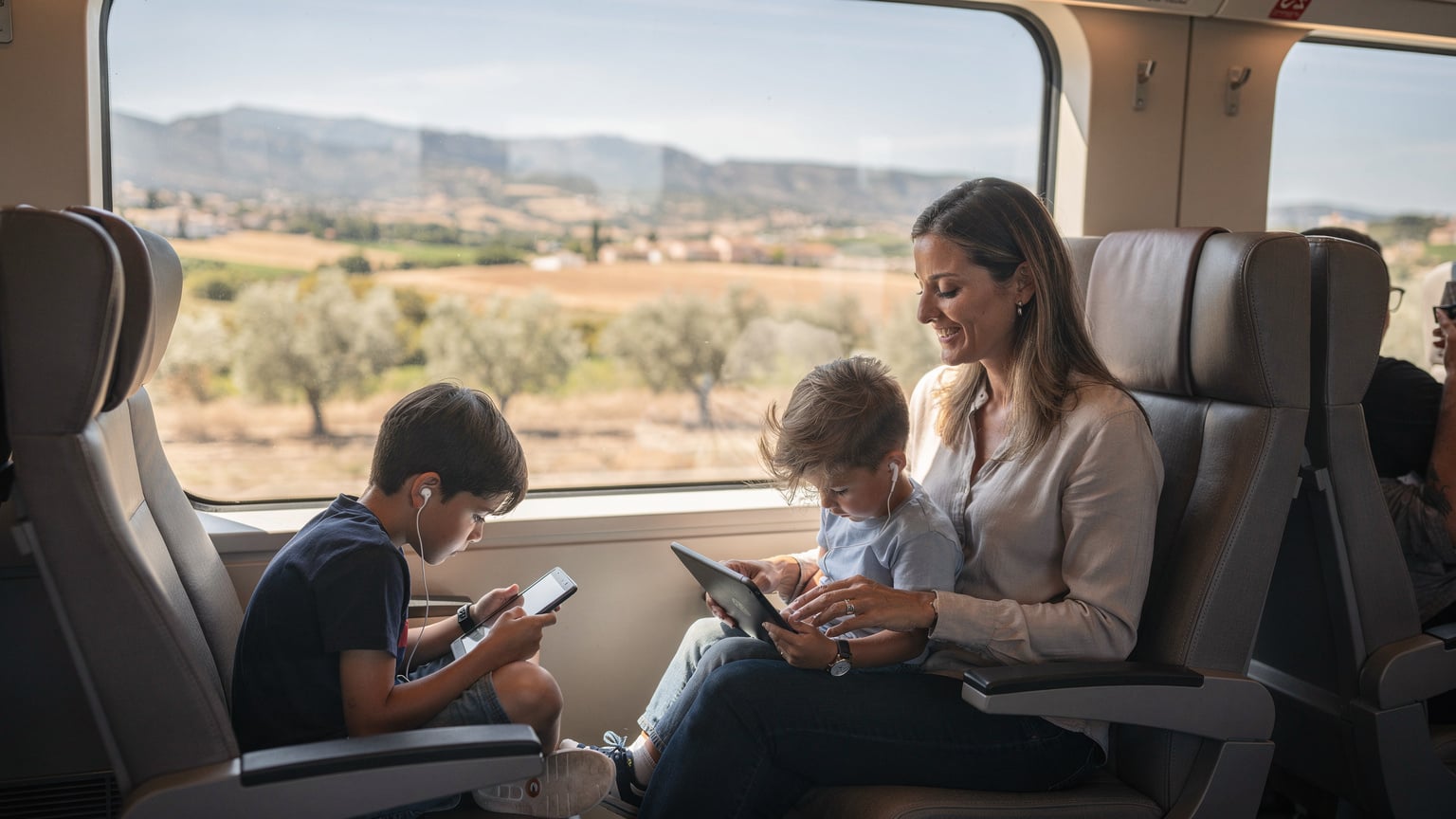 Una madre sonriente y sus dos hijos usando tablets en un tren AVE, disfrutando de la conectividad y el paisaje español.
