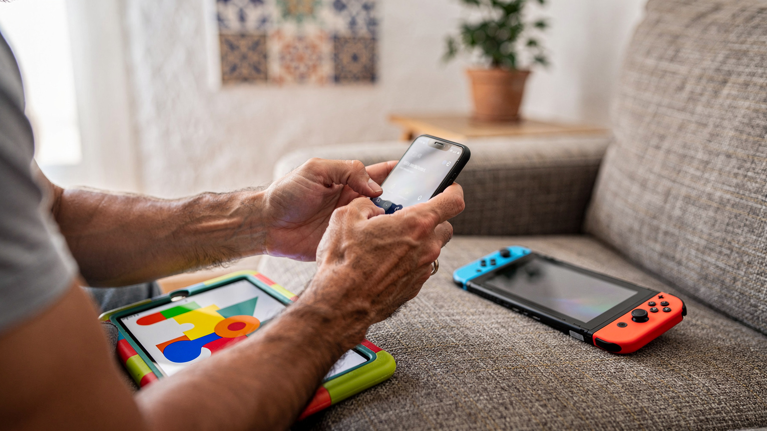 Manos de un padre configurando el hotspot en un smartphone, con un iPad infantil y una Nintendo Switch en un sofá, en un apartamento en Valencia.