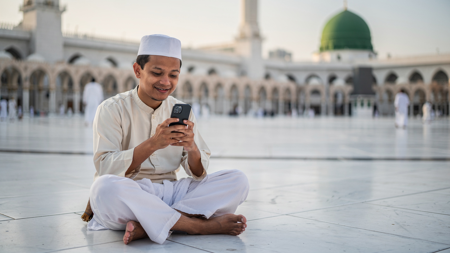 Seorang jemaah lelaki dari Malaysia sedang menggunakan telefon pintarnya di perkarangan Masjid Nabawi yang indah di Madinah, mungkin sedang membuat panggilan video kepada ahli keluarga, dengan kubah hijau di latar belakang.