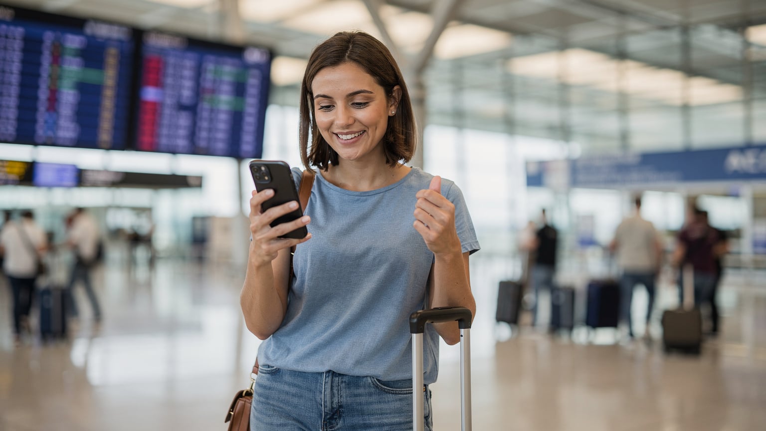 Una giovane donna con un'espressione felice e un bagaglio a mano guarda il suo smartphone, che mostra un'icona di piena connessione di rete, al terminal degli arrivi dell'aeroporto internazionale di Atene, con i tabelloni dei voli sullo sfondo.