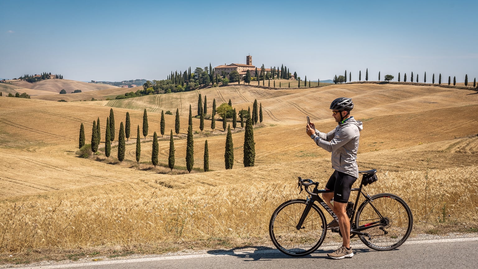 Een fietser maakt een foto van het adembenemende uitzicht over de Val d'Orcia in Toscane, met glooiende heuvels, wijngaarden en iconische cipressen onder een helderblauwe lucht.