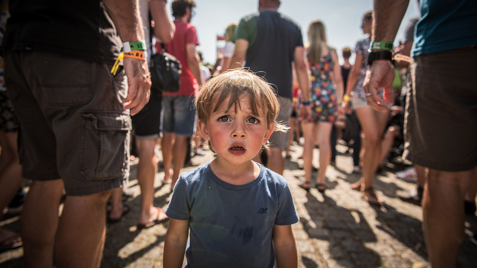 Ein etwa 8-jähriges Kind steht verloren und leicht verängstigt in einer dichten Menschenmenge auf einem Festivalgelände und sucht suchend um sich. Nur erwachsene Beine sind um es herum zu sehen, die ihm die Sicht versperren. Die Szene ist hell und chaotisch, typisch für ein sonniges Outdoor-Festival.