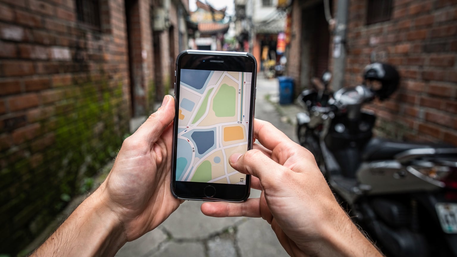 台北の龍山寺近くの路地裏で、スマートフォンで地図アプリを見ながら道を探している旅行者の手元をクローズアップ。周りには古い建物の壁とバイクが写っている。