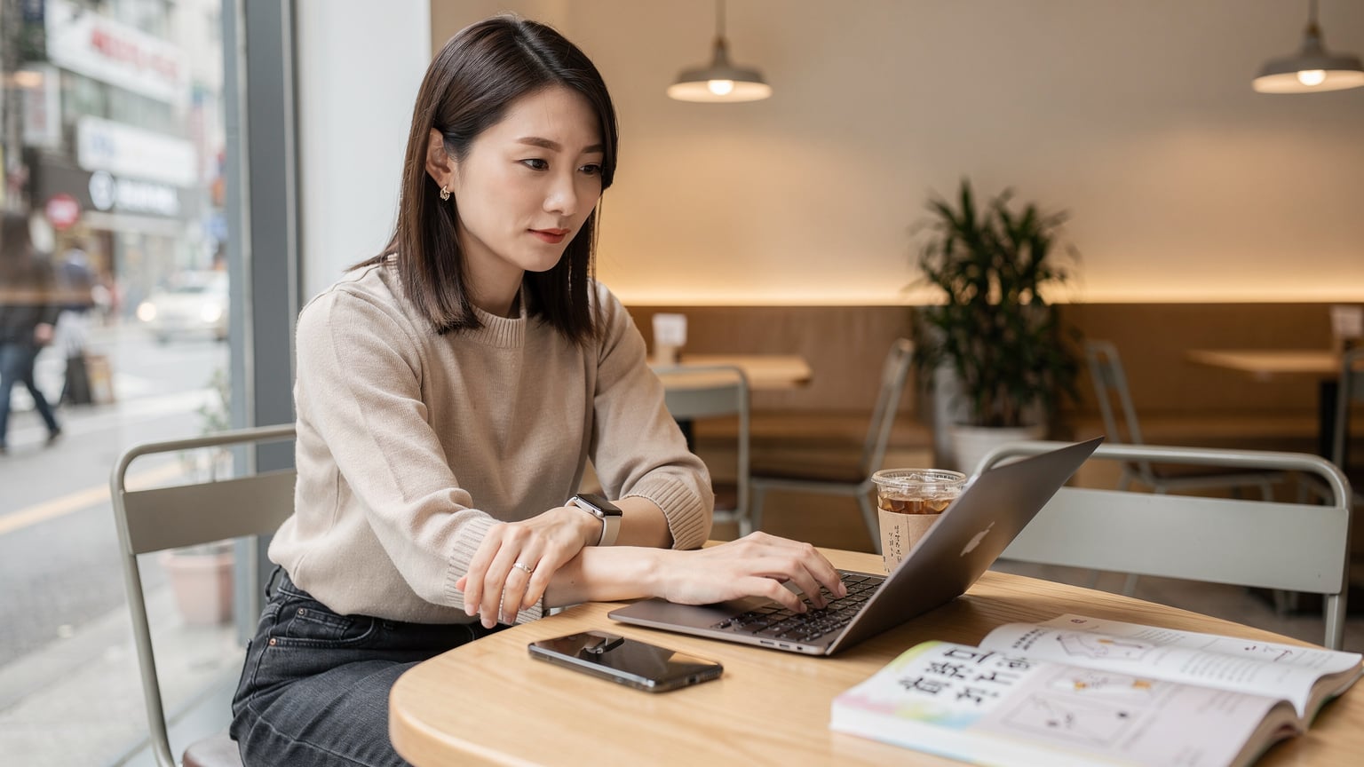 ソウルの弘大にあるカフェで、MacBook AirとiPhoneを使って仕事をしている日本人女性。手元にはアメリカーノと韓国語のガイドブックが置かれている。