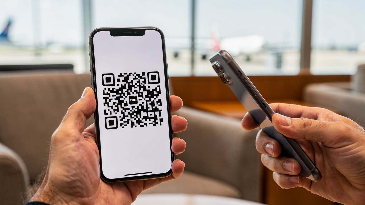 Handen van een reiziger die een QR-code scant op een telefoon om een eSIM te activeren, in een lichte, moderne luchthavenlounge.