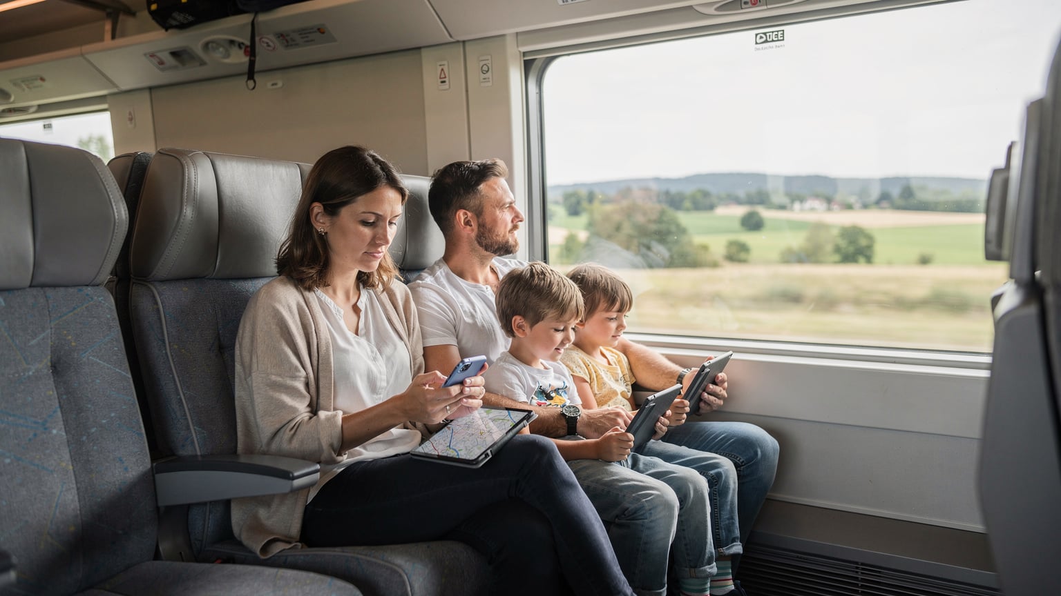 Eine Familie reist mit dem Zug durch Deutschland, Kinder streamen auf Tablets, Eltern nutzen Smartphones.