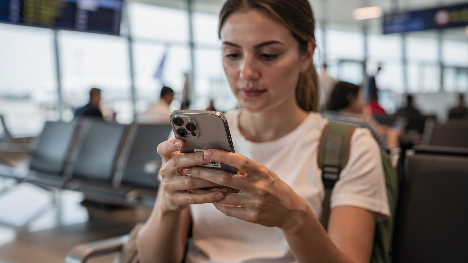 Uma pessoa atenta olhando para o seu iPhone 18, que exibe um código QR para ativação de eSIM, em um ambiente de aeroporto movimentado.
