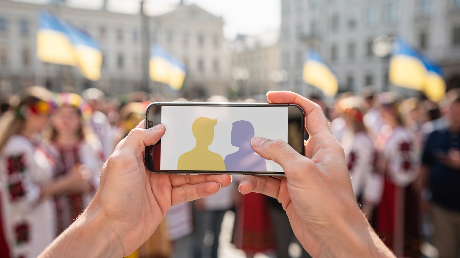 Крупний план рук, що тримають смартфон з відкритим додатком для відеодзвінків, на тлі розмитого святкового заходу з українськими прапорами в Європі.