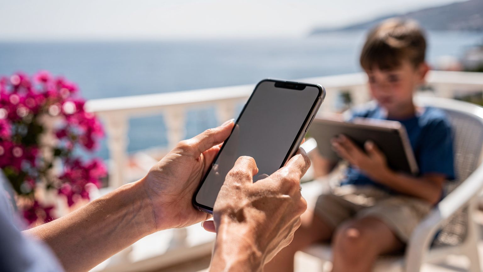 Un primo piano delle mani di una madre mentre attiva la funzione hotspot sul suo smartphone per connettere il tablet del figlio, che è visibile in secondo piano. La scena si svolge su un balcone con vista mare in una località turistica.