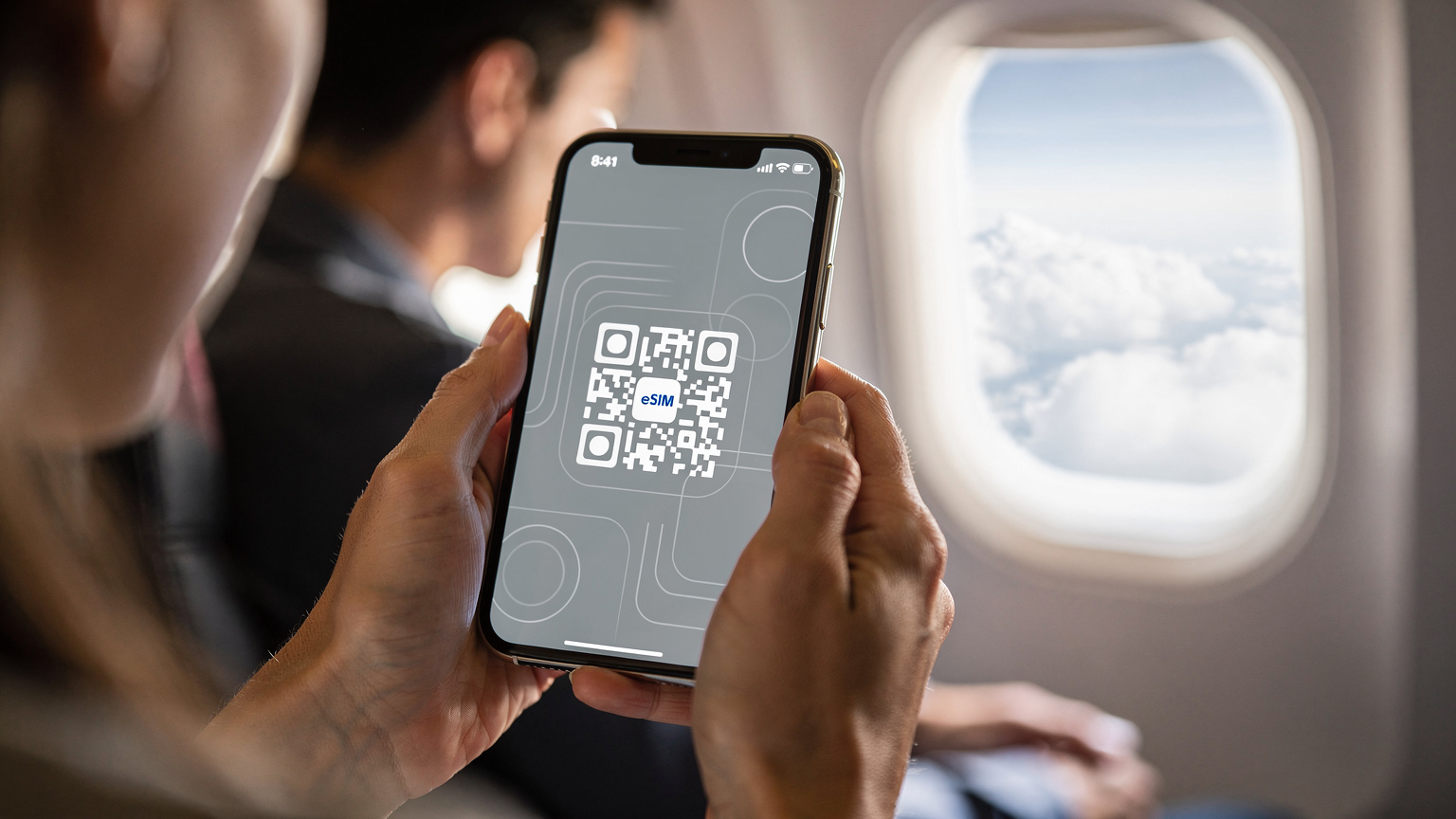 Tangan seorang wanita sedang memegang iPhone dengan layar menampilkan proses instalasi eSIM, memindai QR code. Latar belakang menunjukkan kabin pesawat yang nyaman dengan jendela memperlihatkan awan di luar. Pencahayaan lembut, alami dari jendela pesawat. Difoto dengan lensa makro 100mm, fokus tajam pada tangan dan layar ponsel, dalam gaya Wirecutter product-in-context. Photorealistic, no on-screen text or UI, no readable logos, no superimposed graphics, faces visible but not over-stylised.