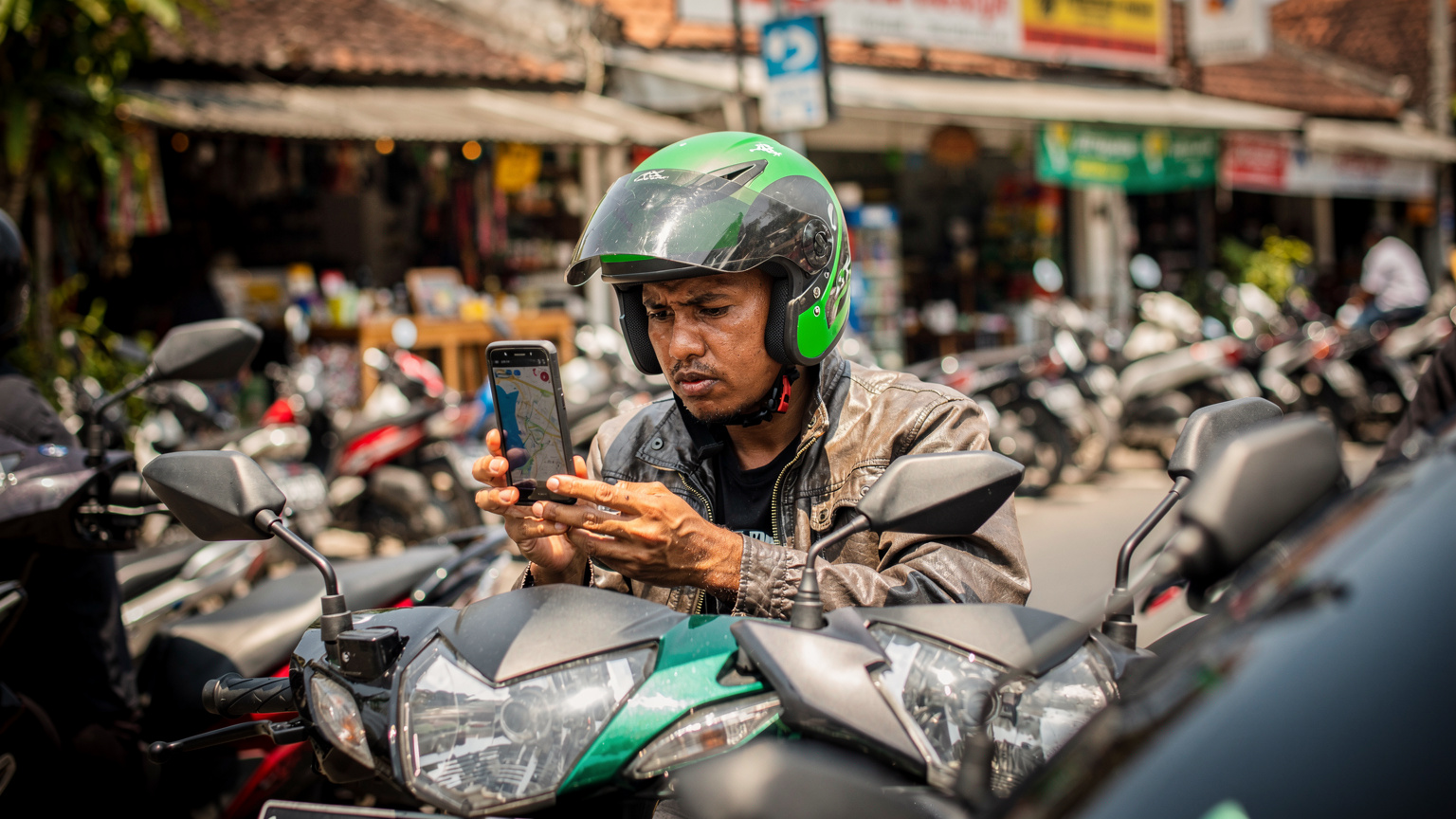 Seorang pengemudi Gojek atau Grab sedang mengamati peta di ponselnya di tengah kemacetan lalu lintas di kawasan Seminyak, Bali, saat siang hari yang cerah. Wajahnya menunjukkan konsentrasi, dengan helm khas Gojek/Grab. Latar belakang menunjukkan deretan toko dan motor yang padat. Sudut pandang jurnalisme jalanan, difoto dengan lensa 50mm, fokus pada pengemudi dan ponselnya. Photorealistic, no on-screen text or UI, no readable logos, no superimposed graphics, faces visible but not over-stylised.