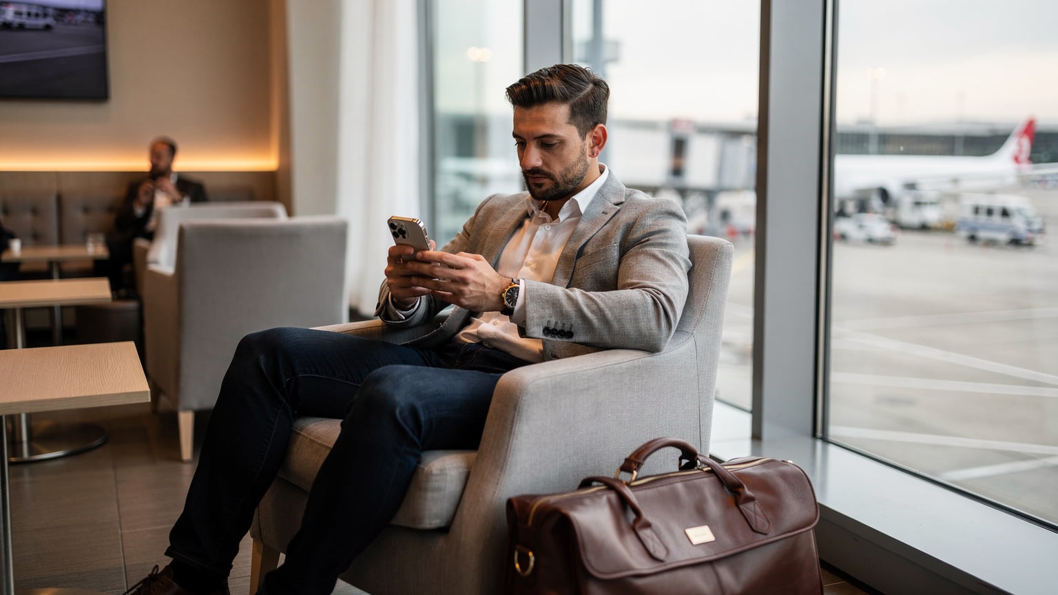 Un viaggiatore seduto in un lounge aeroportuale, con un iPhone 15 in mano, controlla le impostazioni di rete prima del decollo, con il bagaglio a mano accanto.