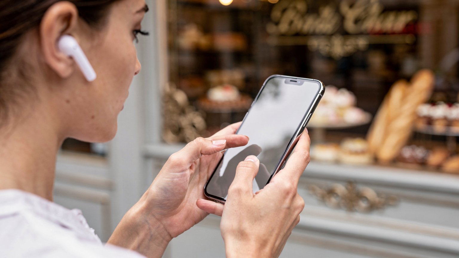 Зблизька жінка в навушниках зосереджено дивиться на екран свого iPhone 17 Pro, на якому відображаються налаштування eSIM, на задньому плані розмитий вітрина паризької пекарні.