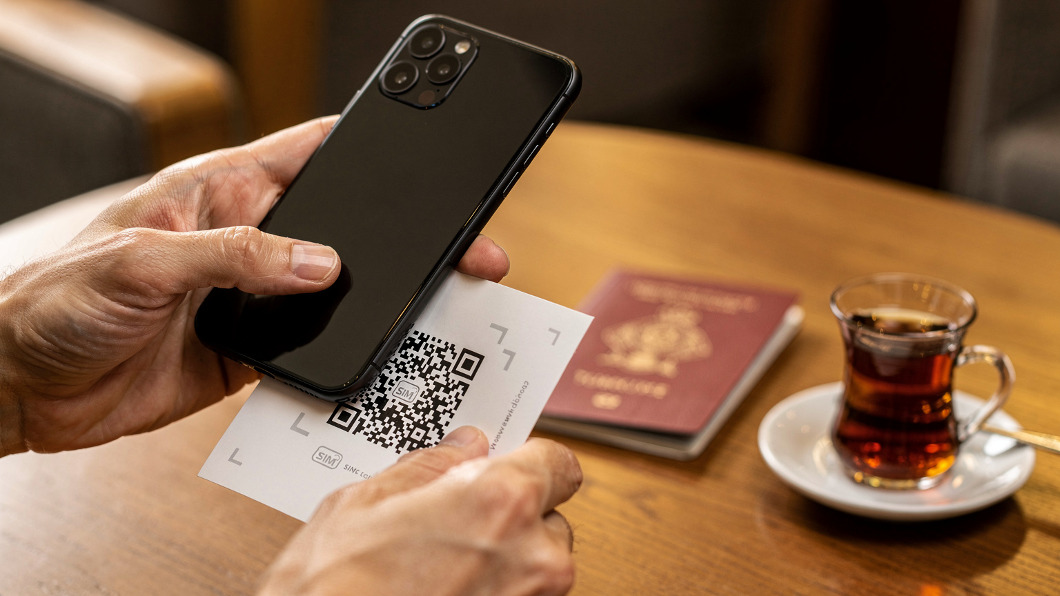 Primer plano de manos de un viajero que escanea un código QR en su teléfono para activar su eSIM, con un pasaporte turco y una taza de té al fondo.
