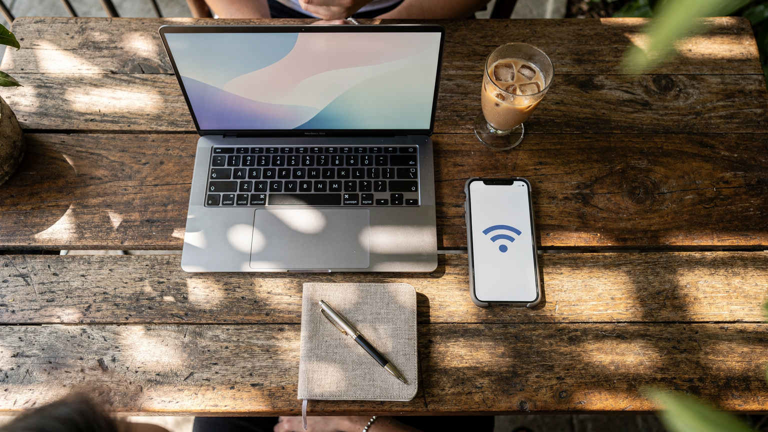 A digital nomad's laptop and smartphone are arranged on a rustic wooden table at a co-working cafe in Bali, with the smartphone actively tethering data to the laptop.