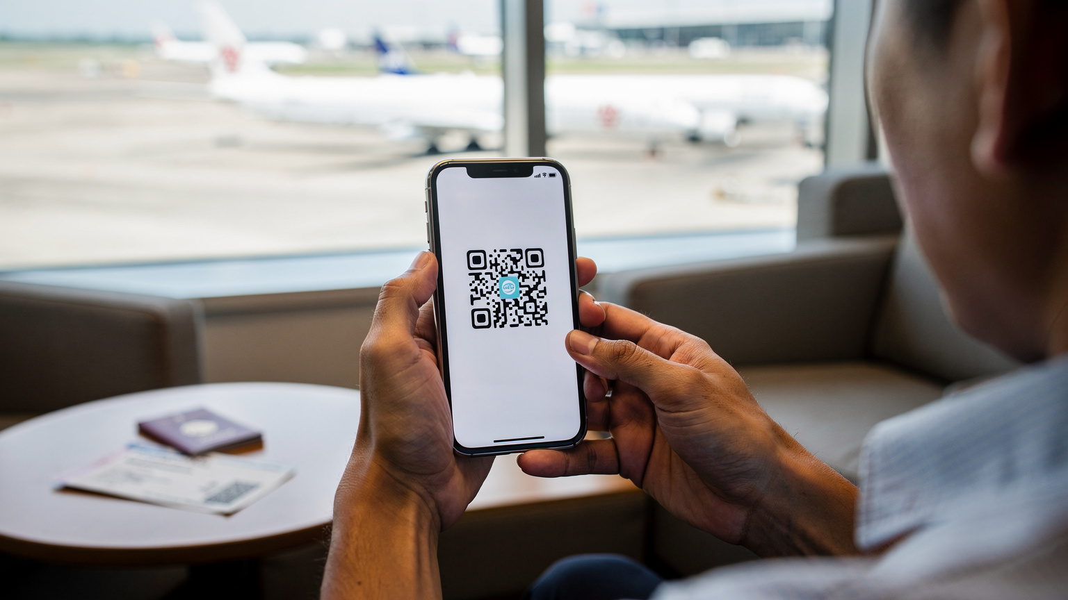 Tangan seorang traveler sedang memegang smartphone dengan tampilan pengaturan seluler, siap untuk memindai kode QR eSIM. Ada paspor dan tiket pesawat di latar belakang.