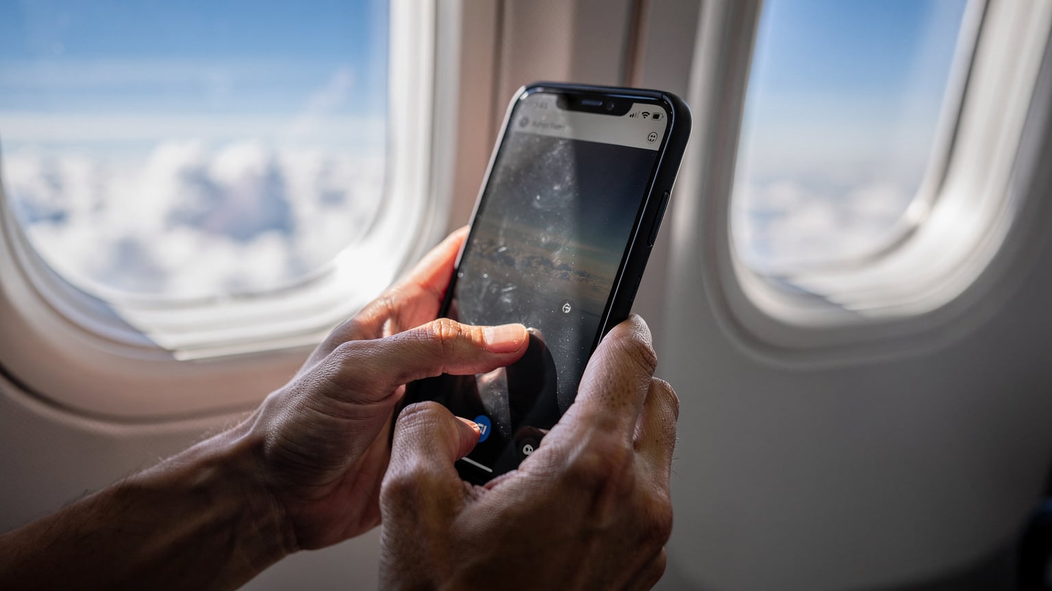 Tangan seseorang sedang mengatur penggunaan data di pengaturan smartphone, dengan latar belakang jendela pesawat, menunjukkan persiapan sebelum mendarat.