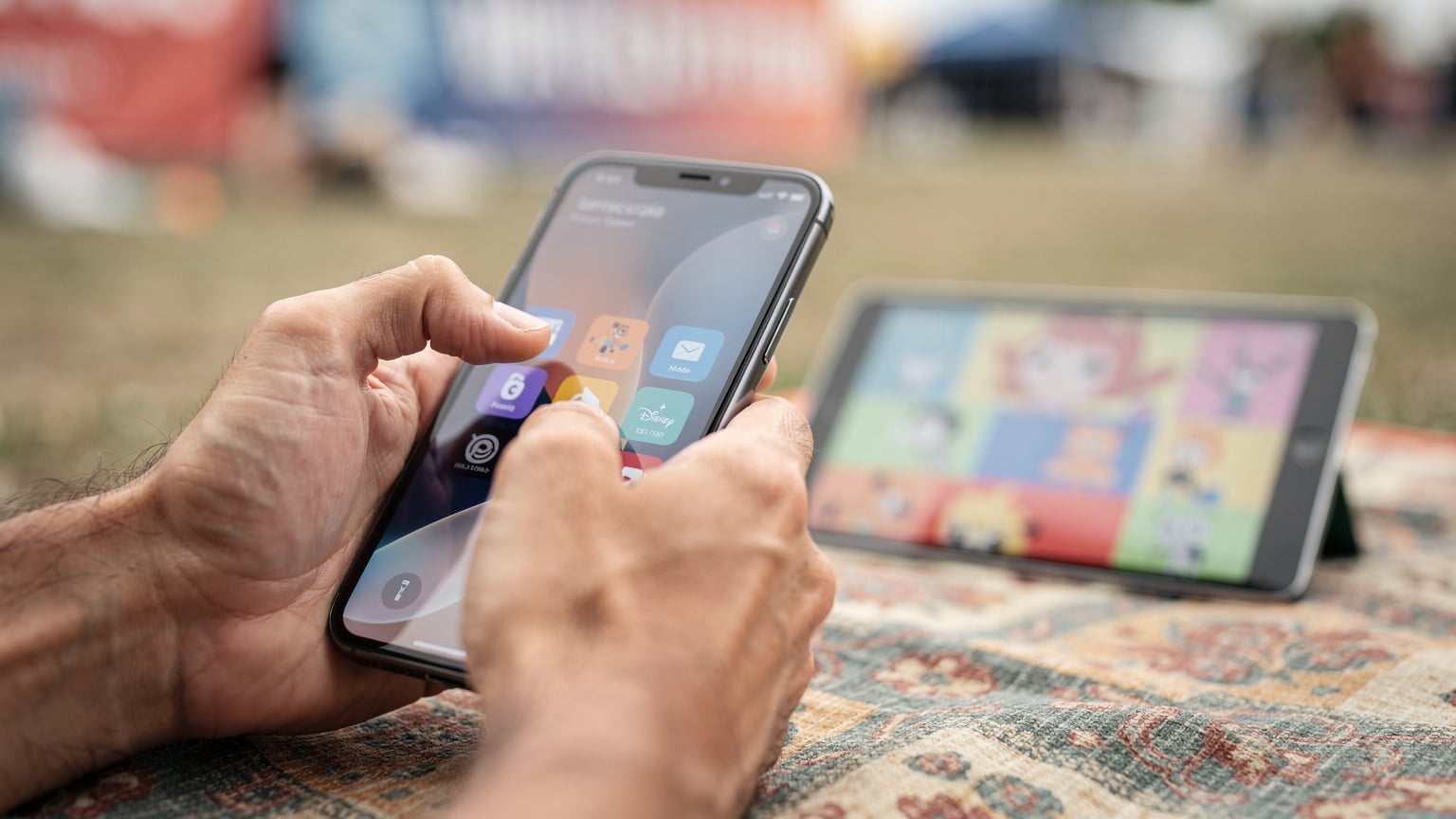 Nærbilde av en hånd som aktiverer hotspot-funksjonen på en smarttelefon, med en iPad i bakgrunnen som viser en strømmetjeneste for barn, på en festival.