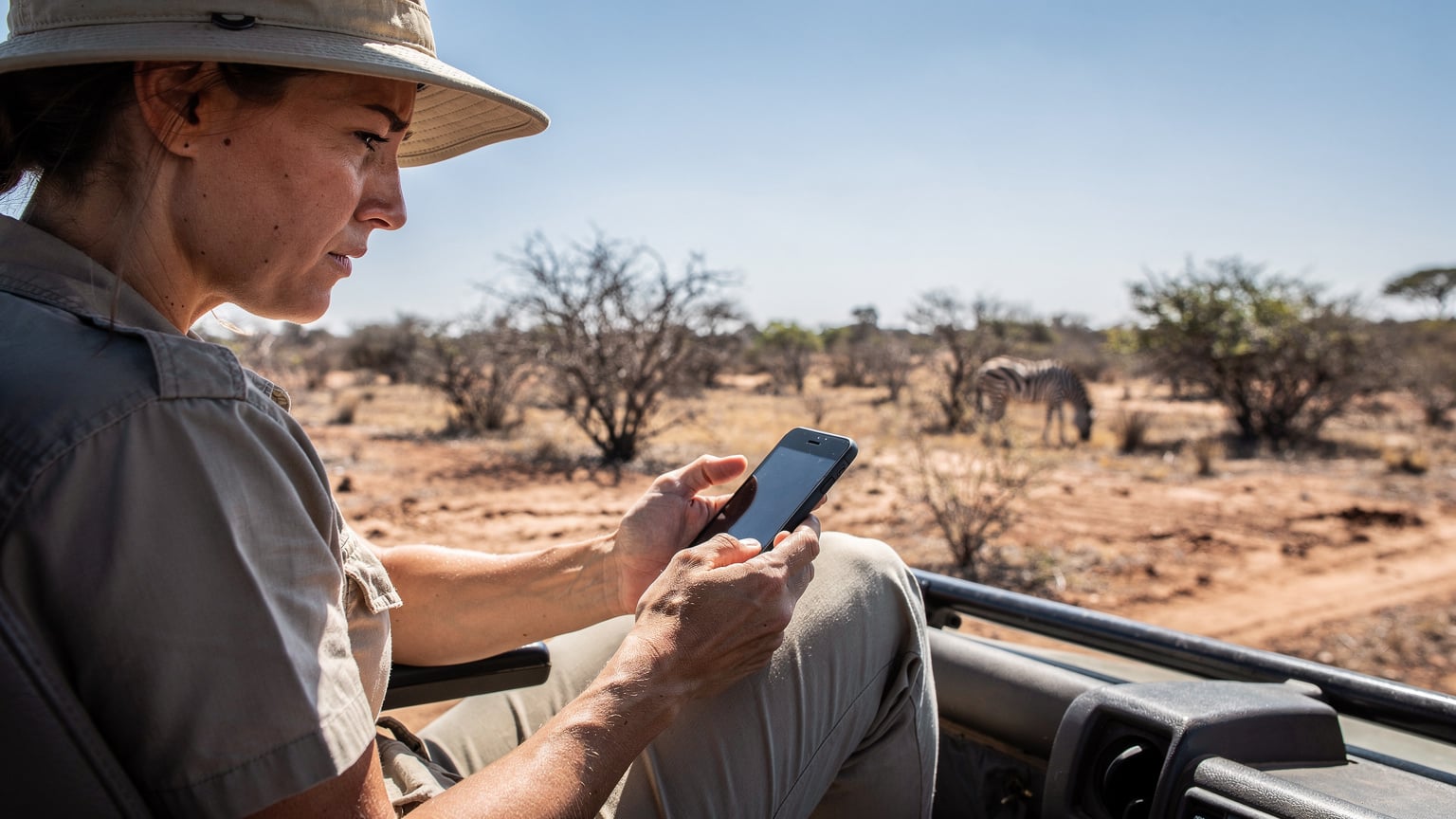 Een vrouw met een hoed kijkt gefrustreerd naar haar smartphone, die 'geen service' toont, terwijl ze in een safari-jeep zit in een afgelegen deel van Kruger National Park, omringd door droge bush en een zebra in de verte.