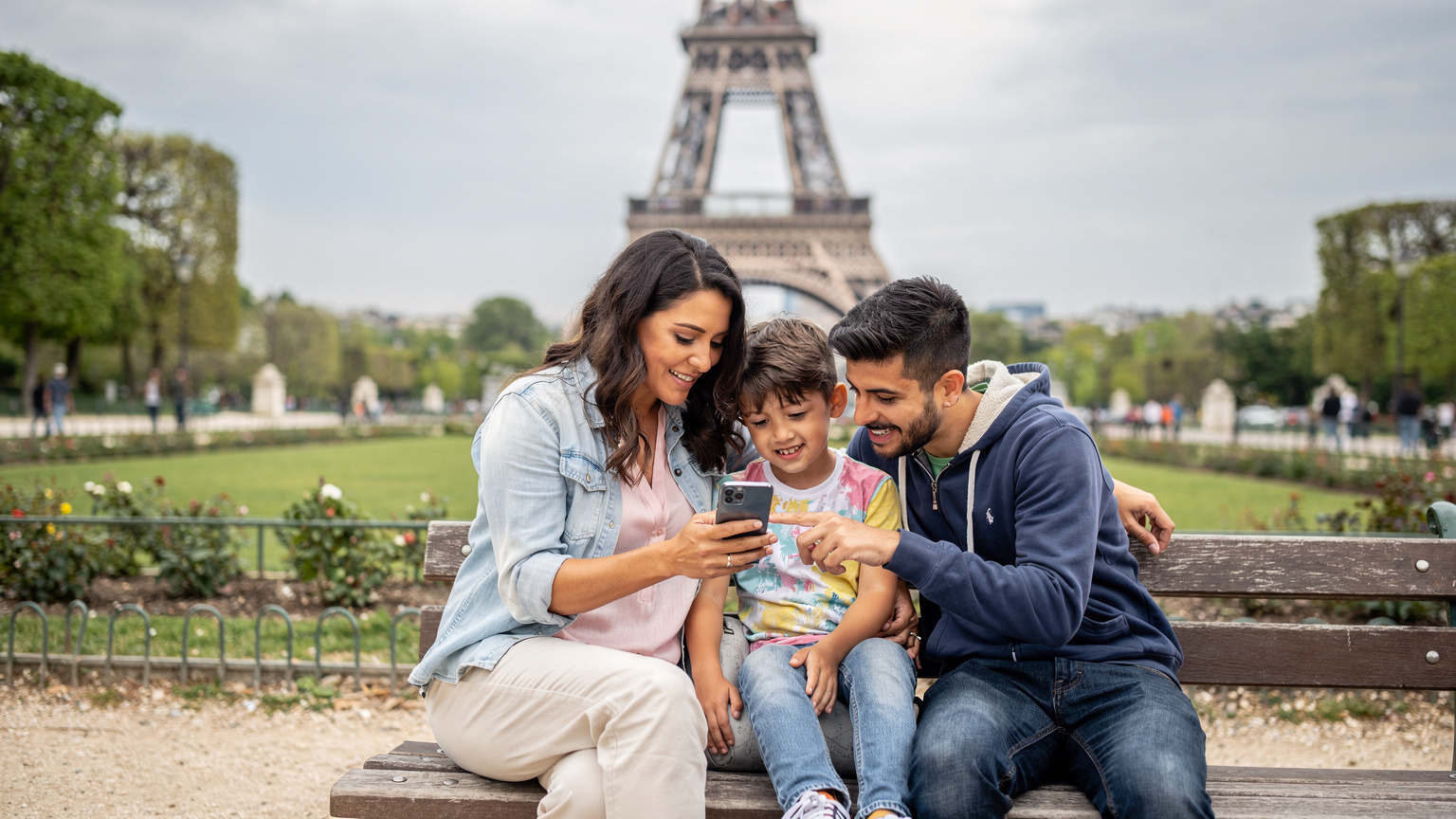 Uma família feliz, pai, mãe e duas crianças, sentados em um banco de parque em frente à Torre Eiffel em Paris, todos olhando para um smartphone com a mãe apontando para a tela, enquanto as crianças observam curiosas.