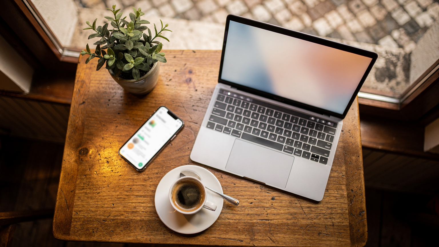 Uma mesa de café em Lisboa com um laptop aberto, um telemóvel exibindo o aplicativo Revolut, um café expresso e um pequeno vaso de flores, simbolizando a vida de um nômade digital.