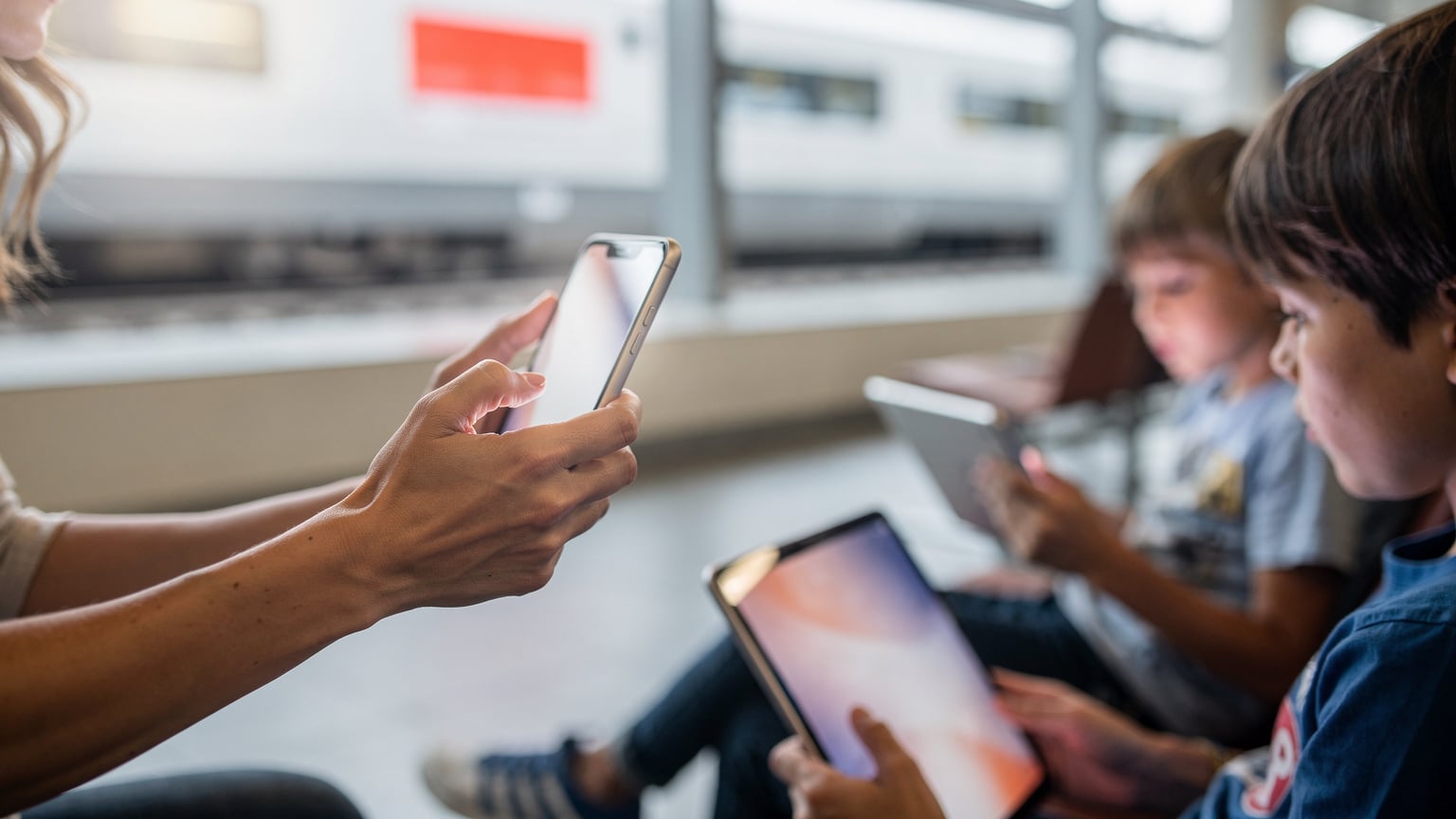 Bir annenin eli, Münih'teki bir tren istasyonunun bekleme salonunda, telefonunda bir mobil hotspot ayarını açıyor. Yanında oturan iki çocuğu, tabletlerinde sakin bir şekilde oyun oynuyor. Arka planda Alman demiryollarının işareti görünüyor.