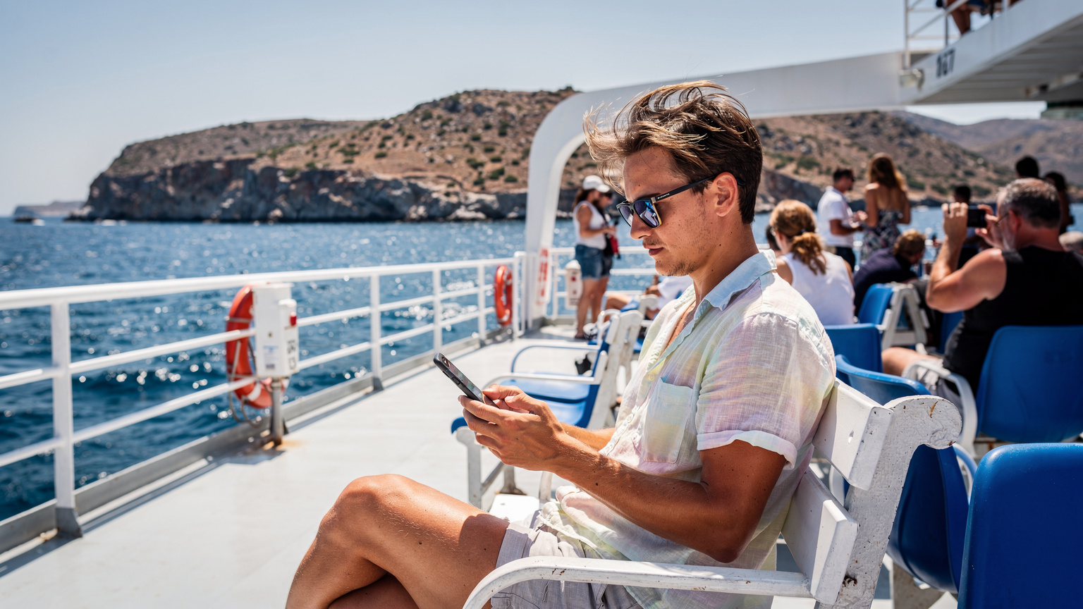 En reisende sjekker sin smarttelefon på en travel ferge som krysser mellom greske øyer, med det blå Egeerhavet og fjerne øylandskap i bakgrunnen.