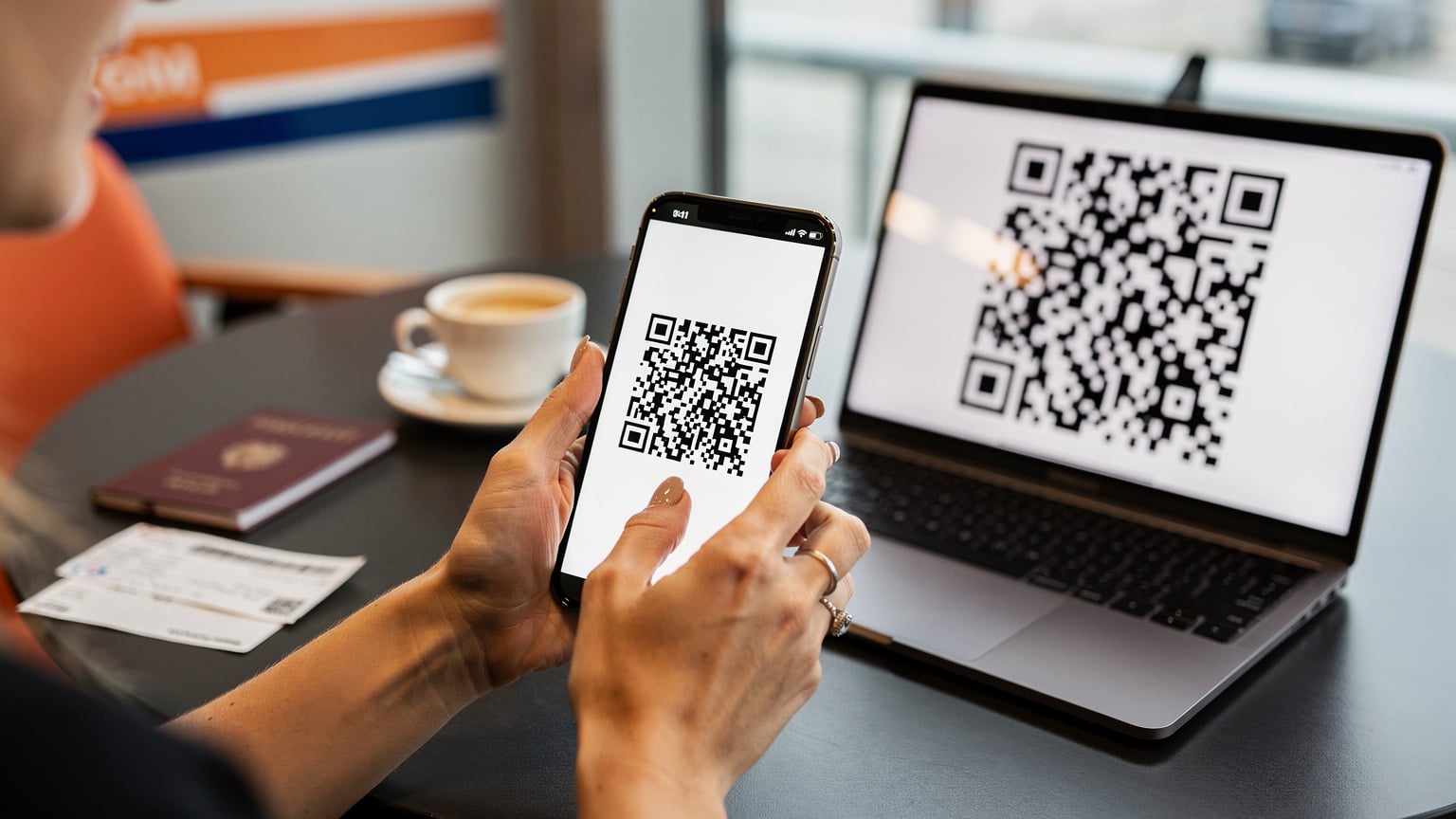 Een close-up van een hand die een iPhone 18 vasthoudt en een QR-code scant van een laptop-scherm, met een paspoort en een boardingpass op een tafelblad, in een café op Schiphol.